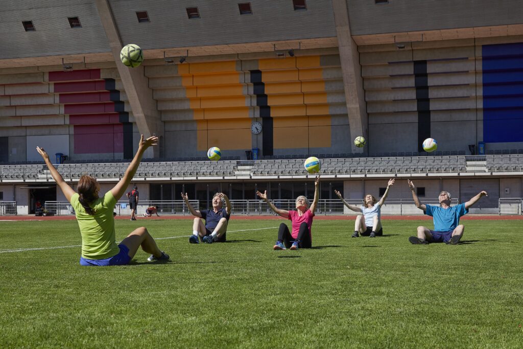 Fünf Frauen und Männer sitzen im Rasen und werfen einen Ball in die Luft.