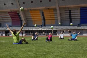 Fünf Frauen und Männer sitzen im Rasen und werfen einen Ball in die Luft.